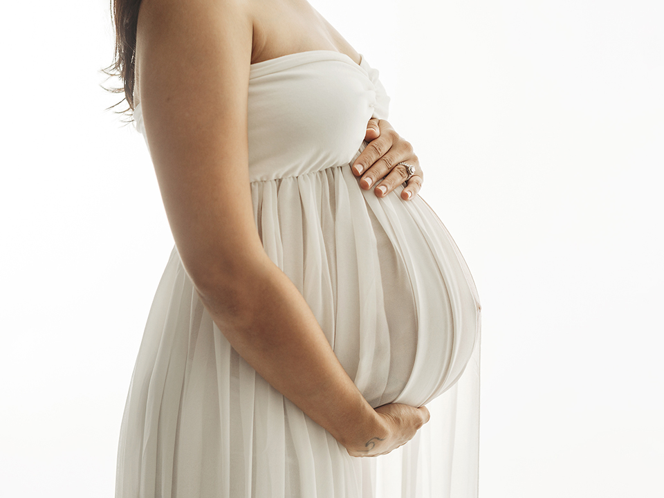 A professional studio photograph of a pregnant woman in a white dress, holding her belly